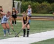 Girls playing field hockey compete for the ball during a match on a lush green sports field.