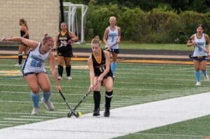 Girls playing field hockey compete for the ball during a match on a lush green sports field.