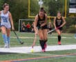 Field hockey players competing on a turf field, focused on the ball, wearing blue and black uniforms.