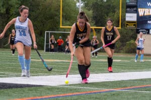 Field hockey players competing on a turf field, focused on the ball, wearing blue and black uniforms.
