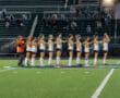 Field hockey team lines up before the game on the field, wearing uniforms, with crowd in the background.