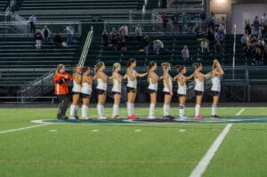 Field hockey team lines up before the game on the field, wearing uniforms, with crowd in the background.