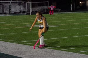 Field hockey player in action on a turf field at night, wearing a Tigers jersey and pink shoes, with goalie in pink gear.