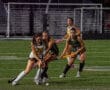 Girls playing competitive field hockey match under stadium lights.