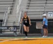 Female field hockey player in action on the field with coach watching in sunny stadium setting.