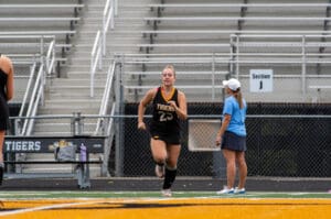 Female field hockey player running on track during practice near coach at sports stadium.