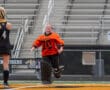 Field hockey goalie running on the field, wearing an orange jersey, celebrating during a game.