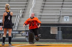 Field hockey goalie running on the field, wearing an orange jersey, celebrating during a game.