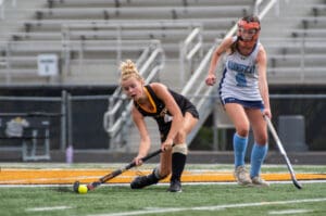 Field hockey players in action, intense match moment on turf. Players wearing black and blue, focused on the ball.
