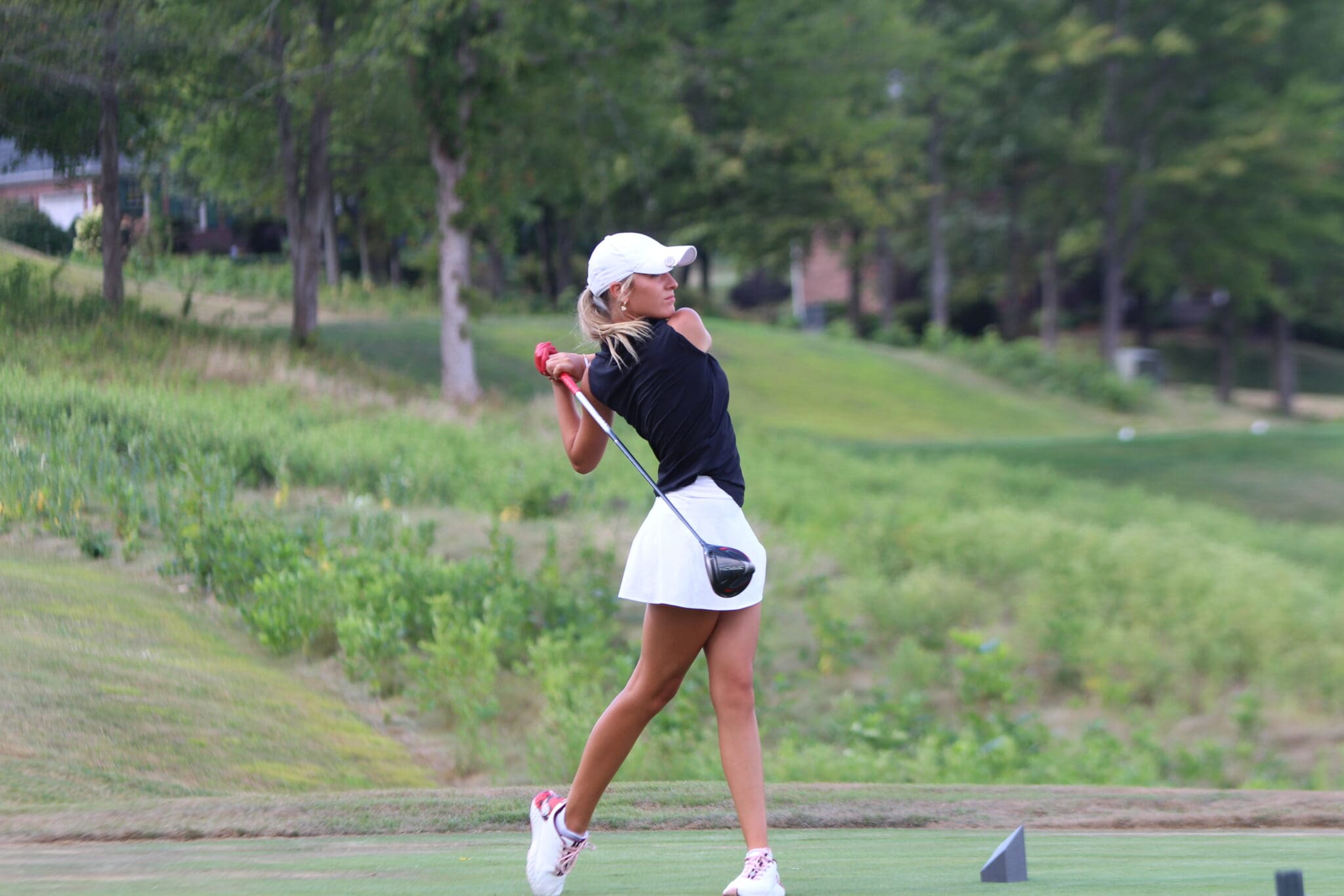 Female golfer in swing motion on a lush golf course, wearing a white skirt and cap, showcasing golfing skills.