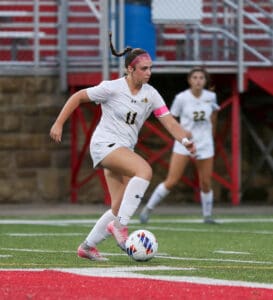 Soccer player wearing white jersey dribbles ball on field, teammate in background.