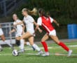 Soccer match action: player in white dribbles past defender in red on a field at dusk.