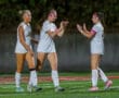 Three female soccer players in white uniforms celebrate on a field, wearing pink armbands and headbands.