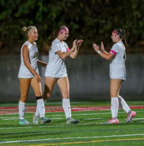 Three female soccer players in white uniforms celebrate on a field, wearing pink armbands and headbands.