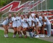 Group of female athletes in soccer uniforms posing together on a sports field, smiling at the camera.