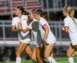 Four female soccer players in white uniforms celebrate on the field during a match.