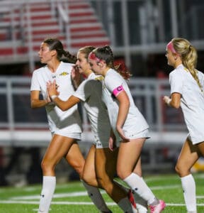 Four female soccer players in white uniforms celebrate on the field during a match.
