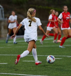 Soccer player in white jersey, number 7, dribbling ball on field, facing opposing players in red uniforms.