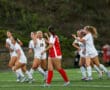 Soccer players in white uniforms celebrating a goal on the field, with an opposing player in red.