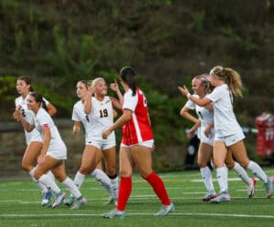 Soccer players in white uniforms celebrating a goal on the field, with an opposing player in red.