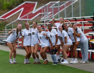 Group of female athletes in soccer uniforms posing together on a sports field, smiling at the camera.