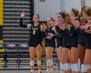 Volleyball player in black Tigers jersey waving, teammates clapping on the side in a gym.