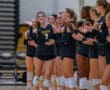Volleyball team players in black uniforms applauding a teammate during a match in a gym.