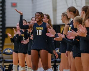 Smiling volleyball player in black jersey claps with teammates in gym, celebrating a point or win.