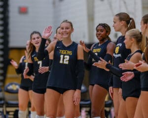 Volleyball team huddle with player number 12 waving and teammates clapping, wearing Tigers jerseys in gym setting.