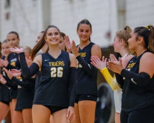 Volleyball players in Tigers jerseys celebrate, smiling and clapping during a match indoors.
