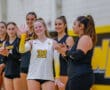 Smiling volleyball player in yellow and black uniform waves while teammates clap in the background.