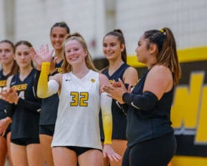 Smiling volleyball player in yellow and black uniform waves while teammates clap in the background.