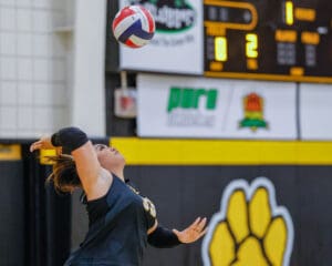 Volleyball player prepares to serve during indoor match, scoreboard in background.