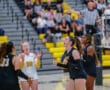 Women's volleyball team celebrates during a match, with blurred spectators in a gymnasium background.