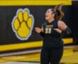 Female basketball player in black uniform, smiling with enthusiasm, stands on court with tiger paw logo in background.