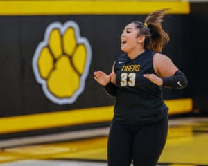 Female basketball player in black uniform, smiling with enthusiasm, stands on court with tiger paw logo in background.