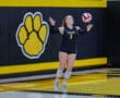 Volleyball player serving during a match, wearing black and yellow jersey, with a paw logo in the background.