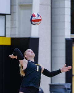 Volleyball player prepares to serve in a gym, focusing on the ball mid-air.