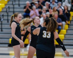 Volleyball players in black uniforms celebrate a point during a match, with teammates and audience cheering in the background.