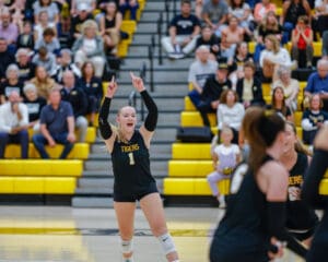Female volleyball player celebrating a point on the court, in front of cheering crowd and yellow bleachers.
