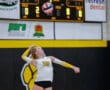 Volleyball player in action serving the ball in a gymnasium with a scoreboard in the background.