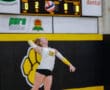 Volleyball player in mid-serve action during a match, wearing yellow and black uniform, gym scoreboard in background.