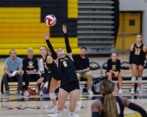 Volleyball player in action, setting ball during a match, teammates and coaches watch intently in background.