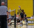 Volleyball player jumps to spike ball over net during competitive match in high school gym.