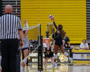 Volleyball player jumps to spike ball over net during competitive match in high school gym.