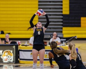 Volleyball player in black jersey setting a ball during a game, with teammates in motion and yellow bleachers in background.