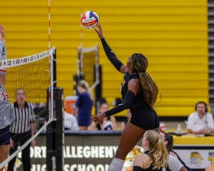 Volleyball player jumps to spike the ball during a high school match in a gymnasium with yellow bleachers.