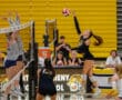 Volleyball player jumps for a spike during a high school match against opponents in gray jerseys.