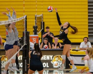 Volleyball player jumps for a spike during a high school match against opponents in gray jerseys.