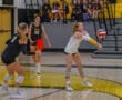 Volleyball player executes a forearm pass during a high school game, with teammates observing in the background.